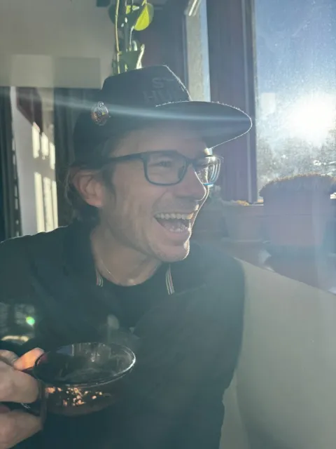 An absolutely dashing white man in glasses and a dark top. He is holding a cup of coffee with visible steam rising, and is wearing a black cap that says stay human.